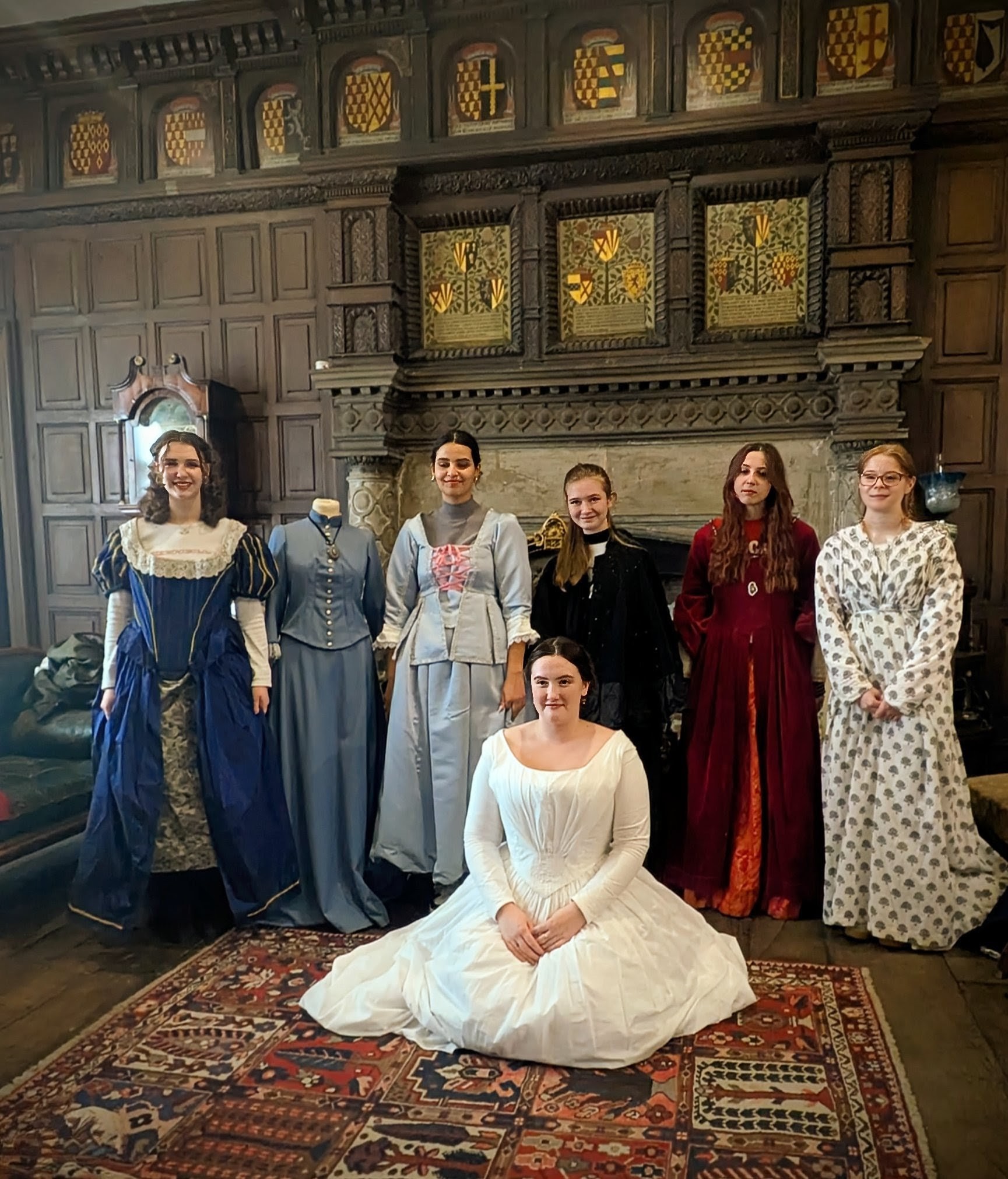 people sitting and standing in Victorian clothes by a fireplace and wooden panel walls
