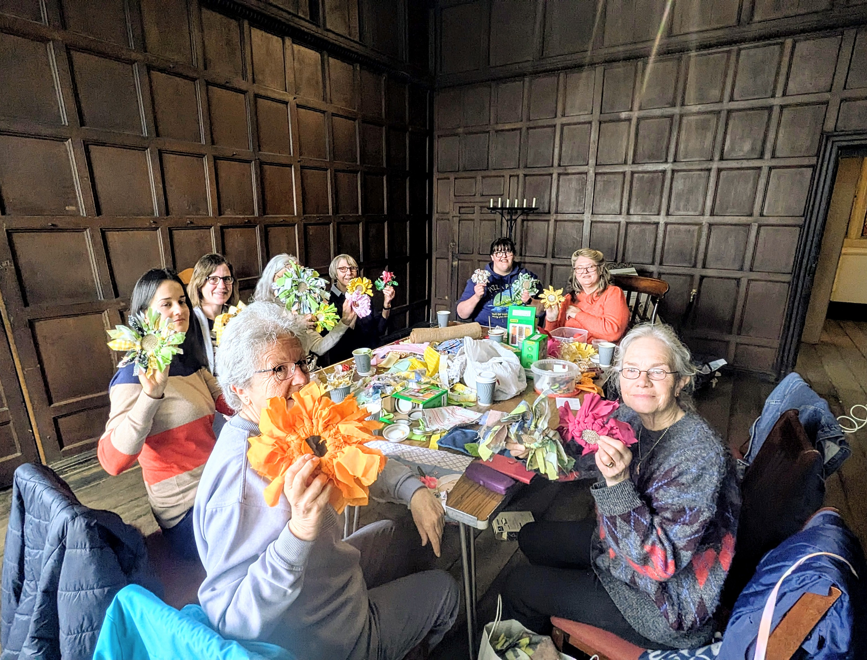 photo of a group of people in a room with dark wooden panelling, holding handmade material and wool flowers