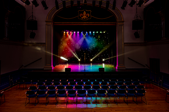 A theatre stage and floor in the dark with rows of chairs and multicoloured lights shining out from the stage