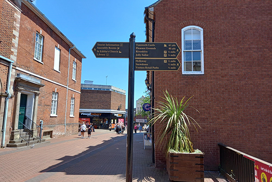 A fingerpost with three signs in the middle of the picture. Two buidings with a pathway in the middle are behind it.