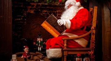 Santa sat on a chair by a fireplace reading a big book
